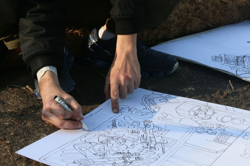 A man in a hoodie uses a black texta to draw a map on a piece of paper