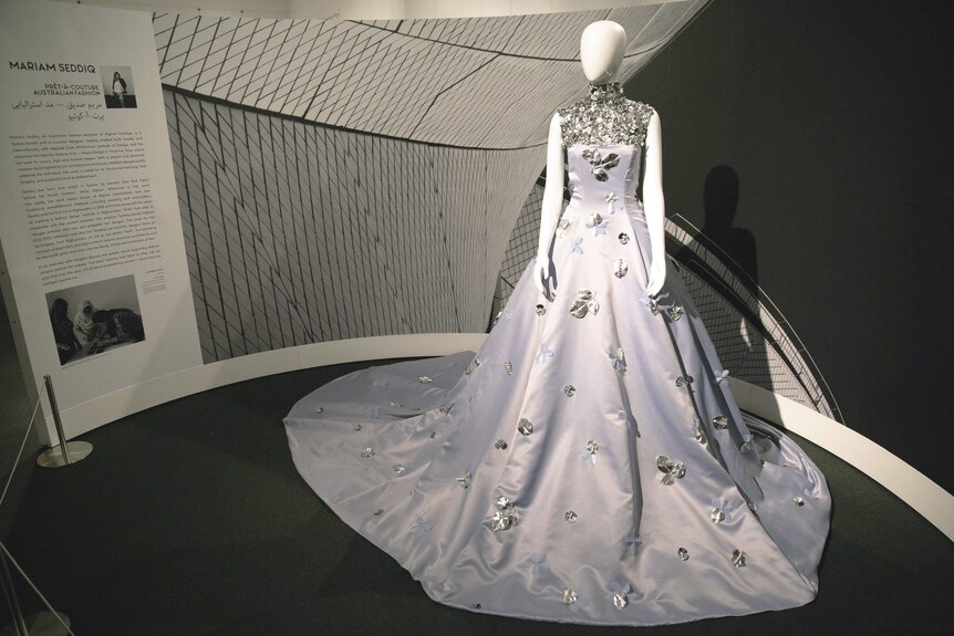 A white gown with elaborate silver embellishments on the bodice and neckline on display in an exhibition.