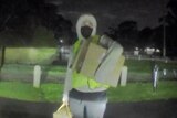 A man wears a grey hoodie with the hood over his head, a yellow vest and gray pants and holds a box under his arm.