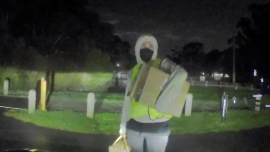 A man wears a grey hoodie with the hood over his head, a yellow vest and gray pants and holds a box under his arm.