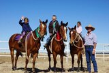 On an outback station, three young kids sit on horses and smile and wave to the camera, their father stands by their side