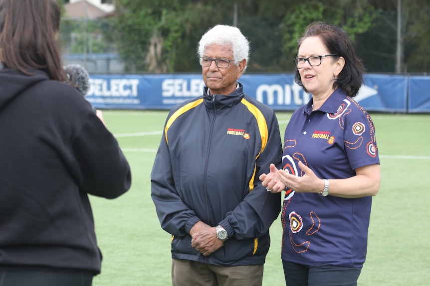 A man and woman speak to a journalist on a football field while being filmed.