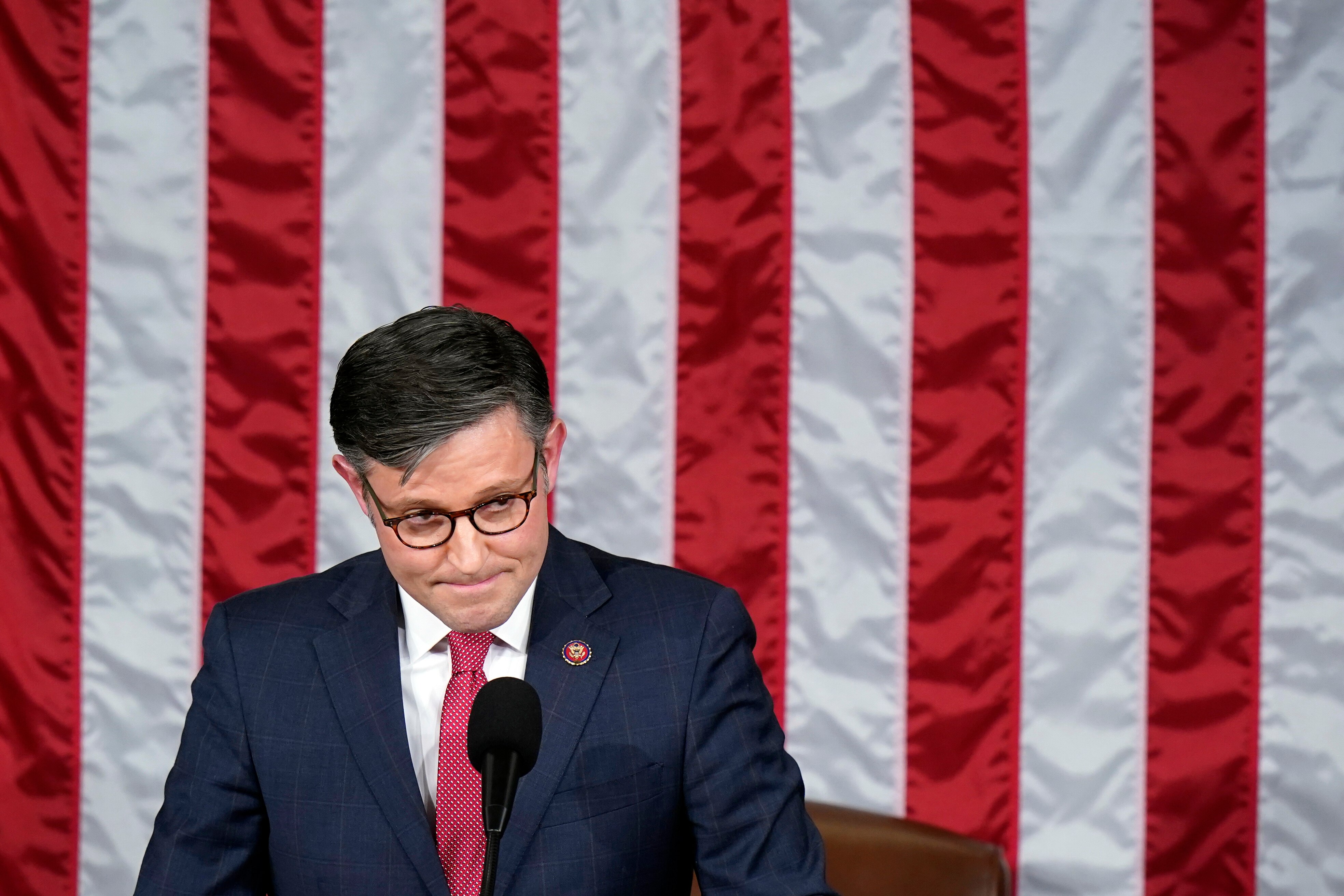 A man standing in front of the US flag wearing a blue suit and red tie and glasses looks pensively to his left