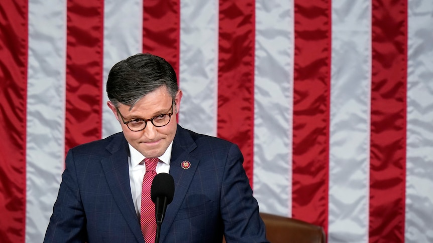 A man standing in front of the US flag wearing a blue suit and red tie and glasses looks pensively to his left