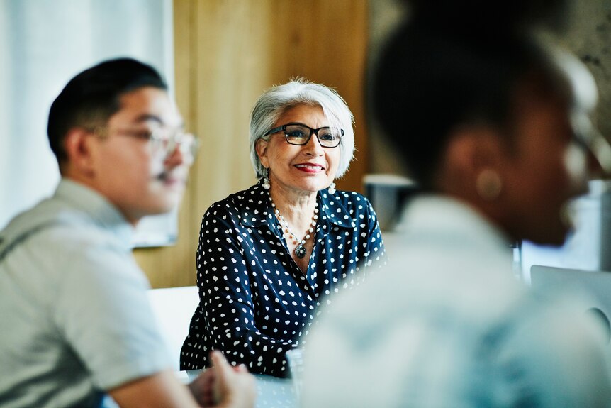 Woman with grey hair and glasses listens to colleagues at a meeting