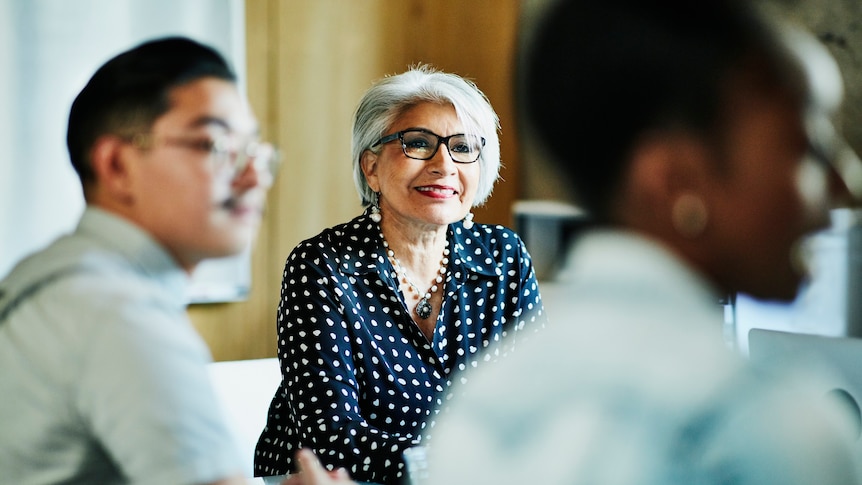 Woman with grey hair and glasses listens to colleagues at a meeting