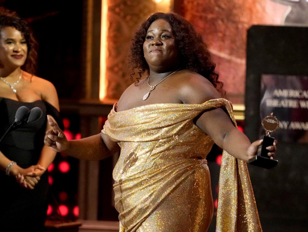 Alex Newell wearing a shiny, gold dress on stage at an awards show 