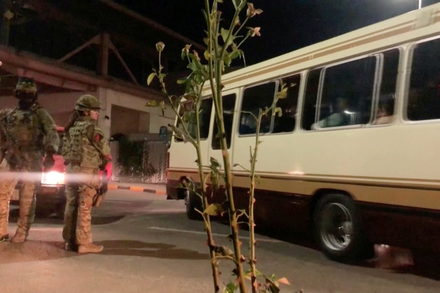 Two men with helmets and fatigues on wait for a bus as it waits behind another vehicle.