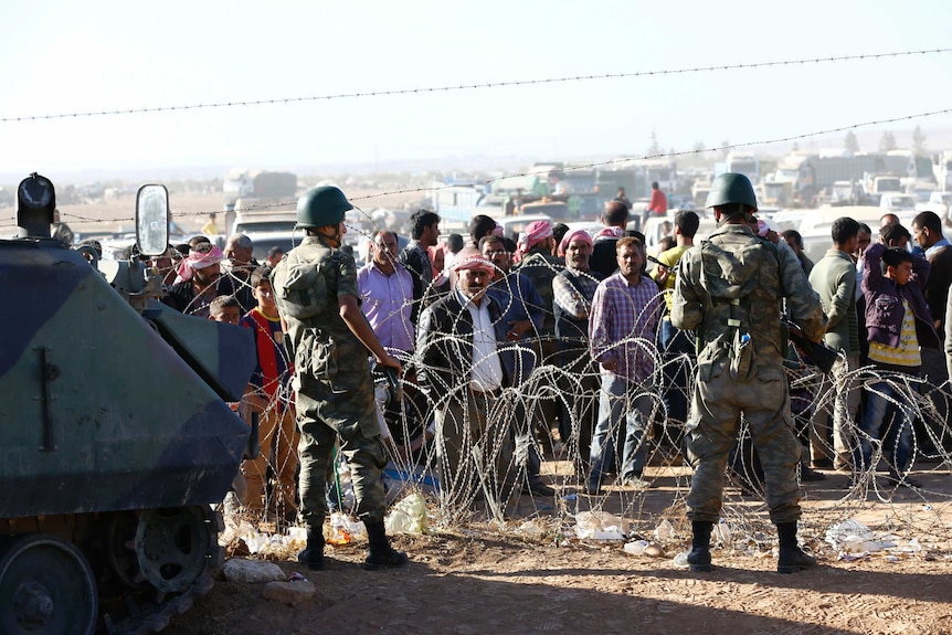 Turkish soldiers stand guard as Syrian Kurds wait behind a border fence.