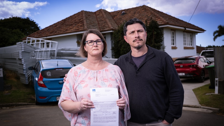 A husband and wife clutching a letter outside a home.