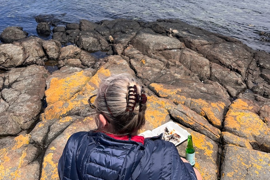 A woman with a box of oysters sits on rocks near the sea