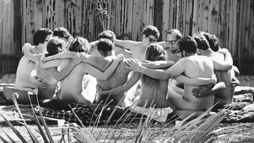 Black and white photo of group of naked men and women sitting in a circle with arms over each others' shoulders.