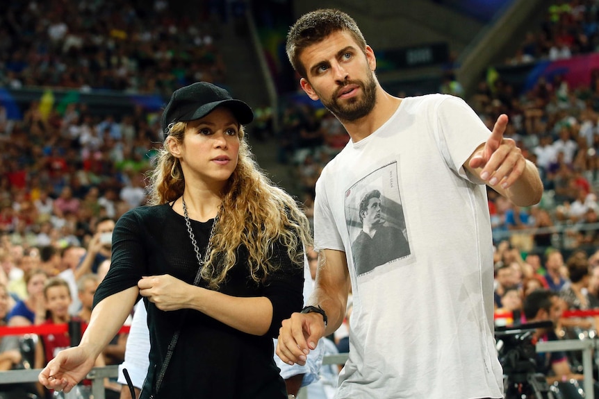 a woman with a black shirt and black baseball cap stands next to a man with a white shirt pointing with an arena crowd behind