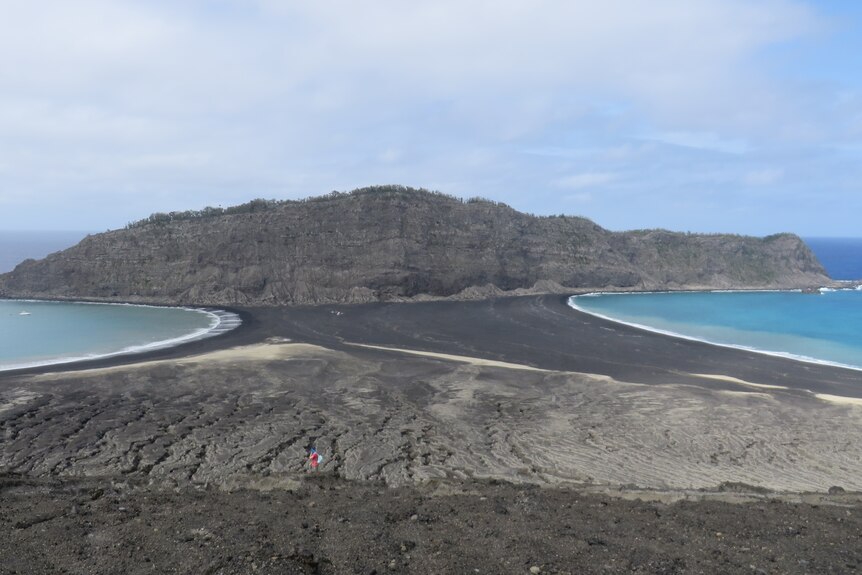A stretch of grey and black volcanic rock runs up to a rugged mound of volcanic rock, with calm seas surrounding. 