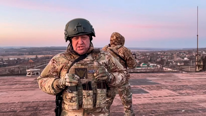 An older white man in heavily armoured combat gear and a helmet stands on a rooftop, with a soldier looking out behind him.