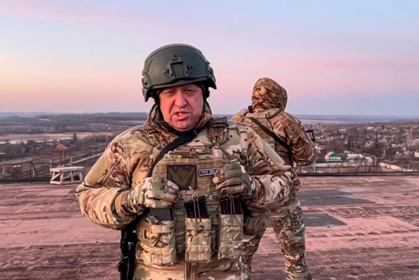 An older white man in heavily armoured combat gear and a helmet stands on a rooftop, with a soldier looking out behind him.