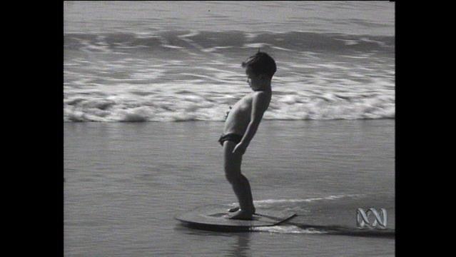 Old photo shows young boy standing on surf board in shallow water