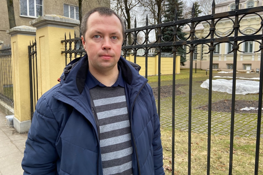Barys, wearing a navy puffer over a striped shirt, looks at the camera. behind him, a black fence surrounds an ornate building