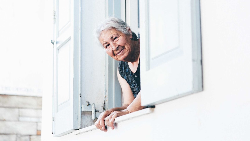 An older woman leans out of a house window and stares into the camera to depict the things we can do to age well.