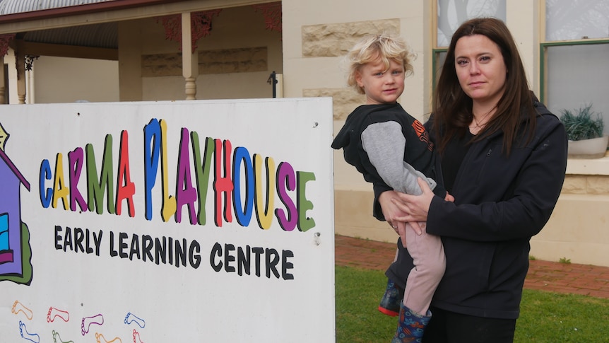 A serious woman in black suit holds a young boy in front of a childcare centre.