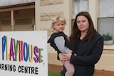 A serious woman in black suit holds a young boy in front of a childcare centre.