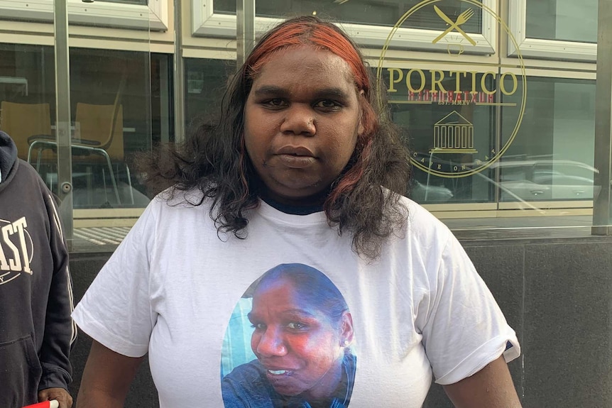 A mid shot of an Aboriginal woman standing outdoors in front of a restaurant wearing a white shirt with a woman's face on it.