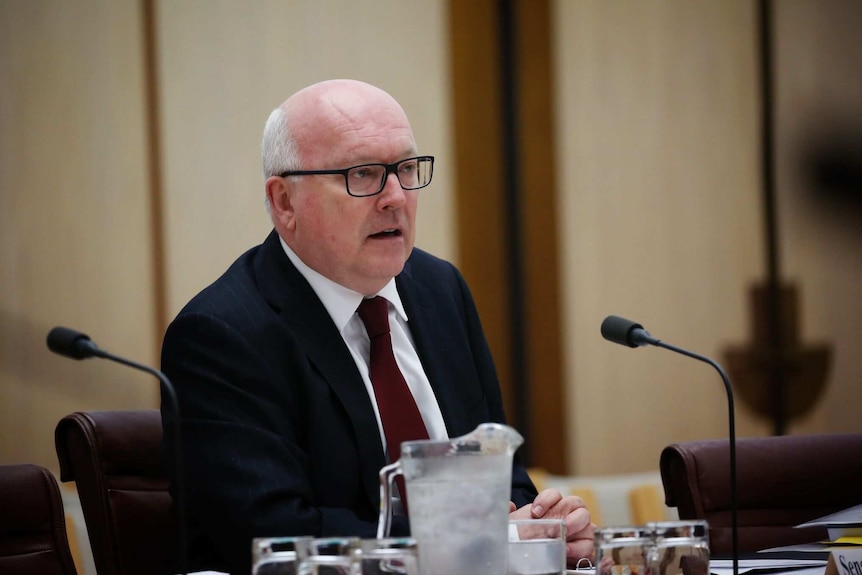 George Brandis sits behind microphones.