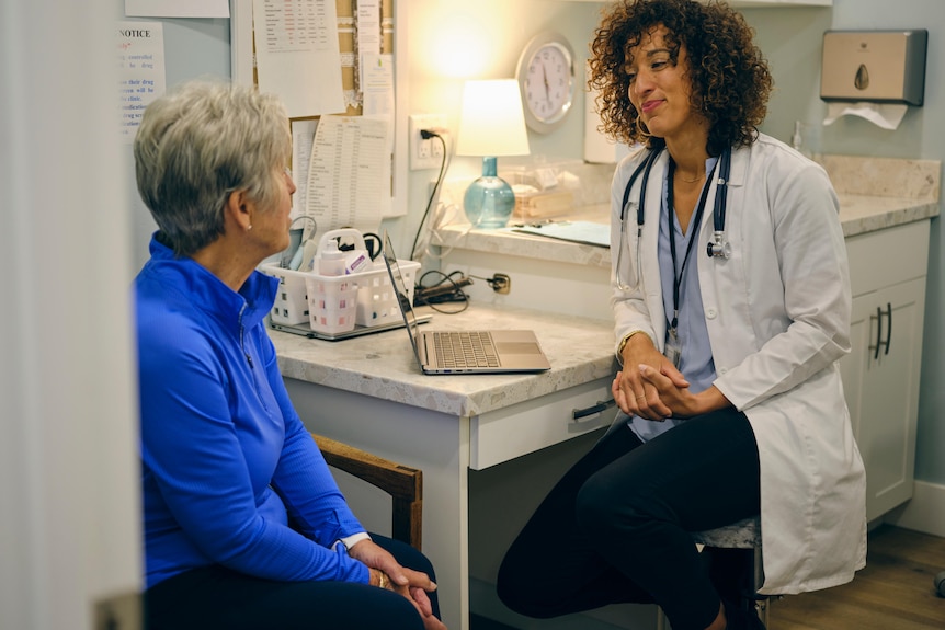 A woman with a blue sweater and grey hair talks to a young female doctor wearing a white coat.