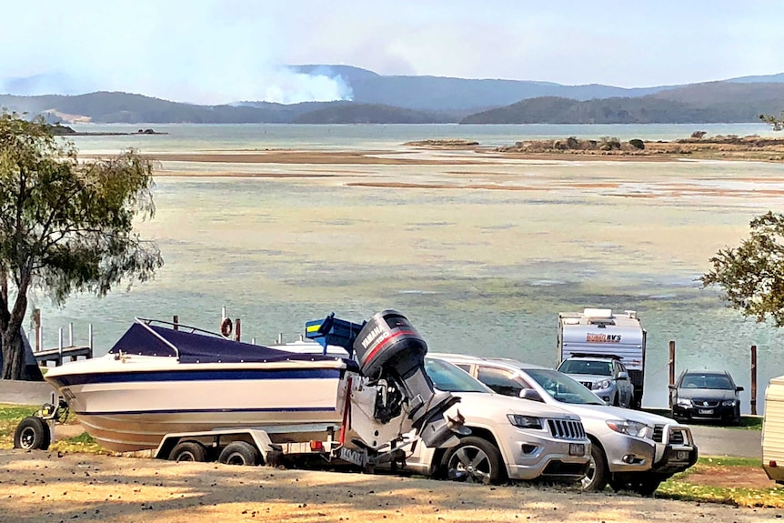 A boat and several cars on the foreshore at Mallacoota with a bushfire burning in the distance.