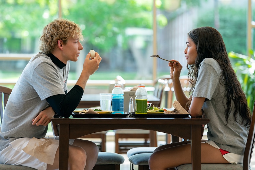 A film still of Mike Faist and Zendaya sitting over a table eating. She is pointing at him with her fork.