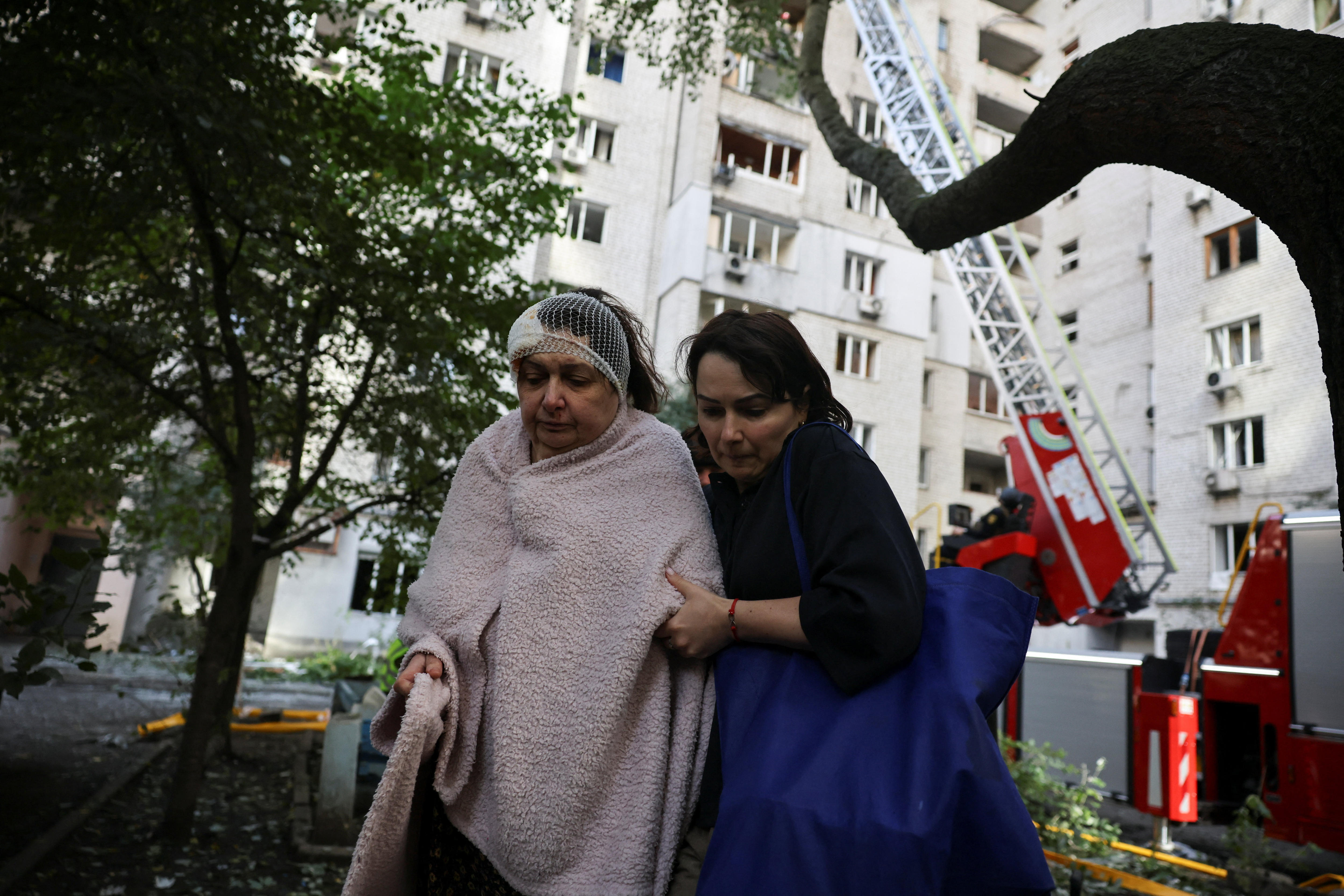 an older woman in a blanket walks away from a damaged building with bandages on her head.