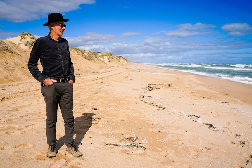 A man looks out to sea from a sandy beach with dunes behind him
