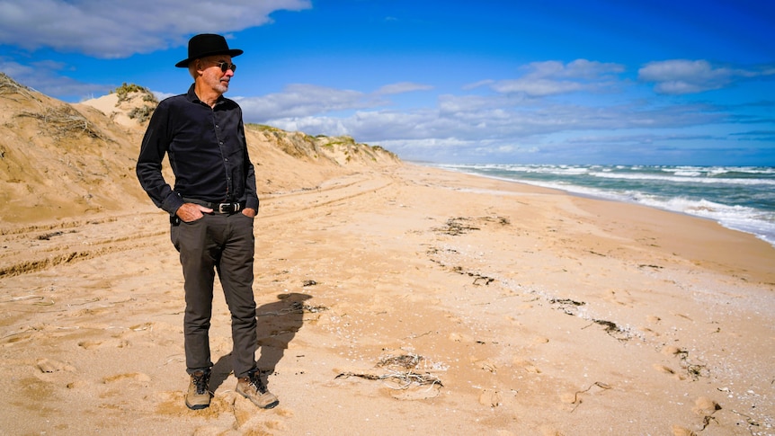 A man looks out to sea from a sandy beach with dunes behind him