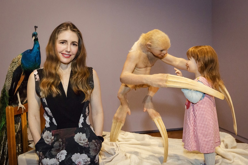 Woman with long hair standing in front of child's bed with a peacock, a genetically engineered "creature" and small child on it.