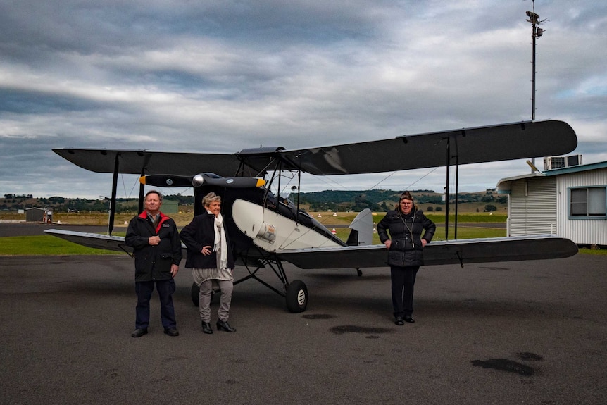 Two men and a woman stand pose in front of a Tiger Moth bi-place on a modern airfield