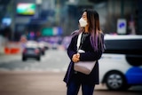 A woman in a face mask with long purple hair stands on a street corner