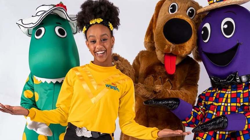 A woman with dark afro hair wearing a yellow top standing with dorothy the dinosaur, wags the dog and henry the octapus. 