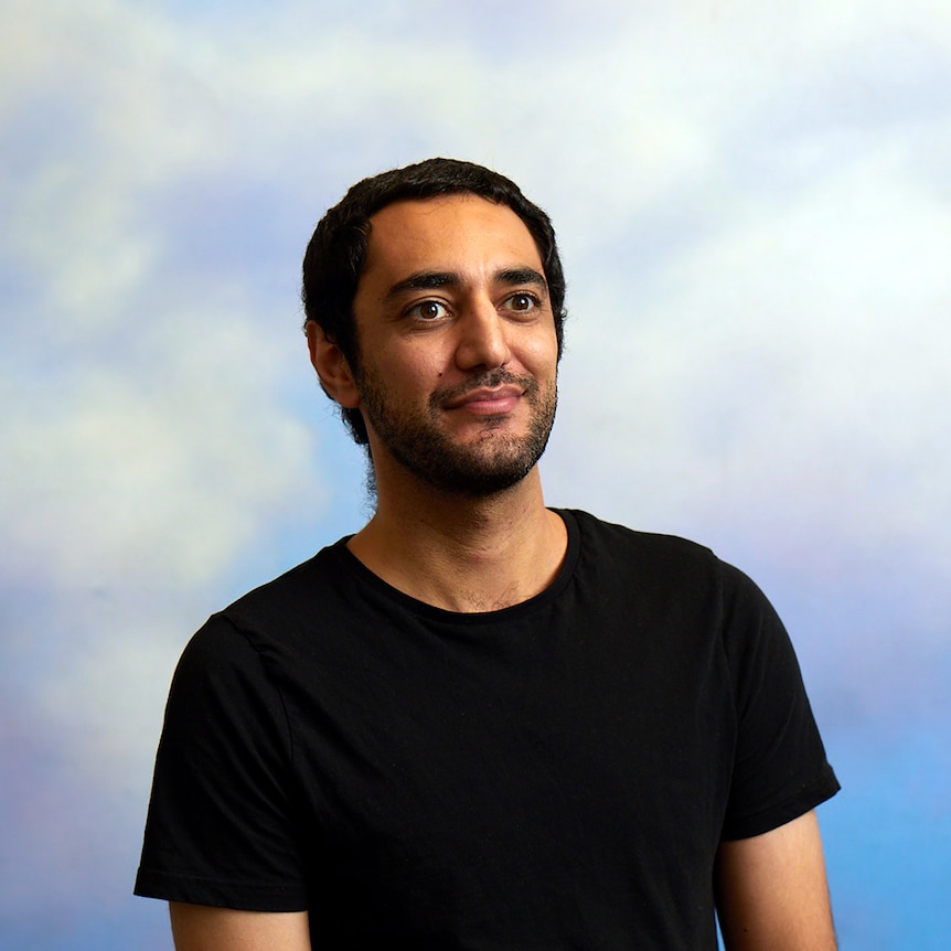 Head and shoulders portrait of a man in a black shirt standing in front of a blue backdrop.