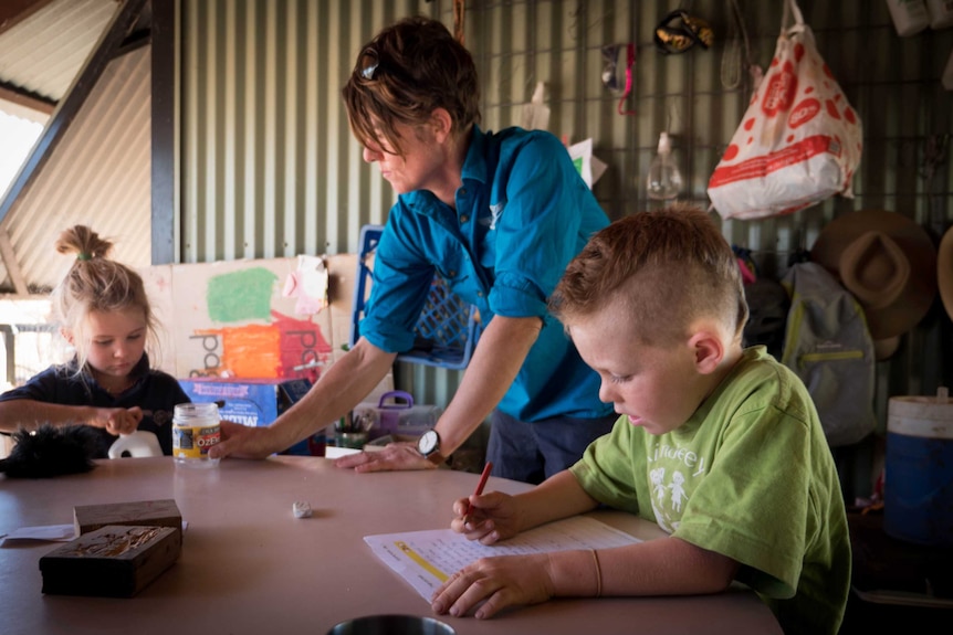 A woman in a blue shirt works with her two children at a table outside.