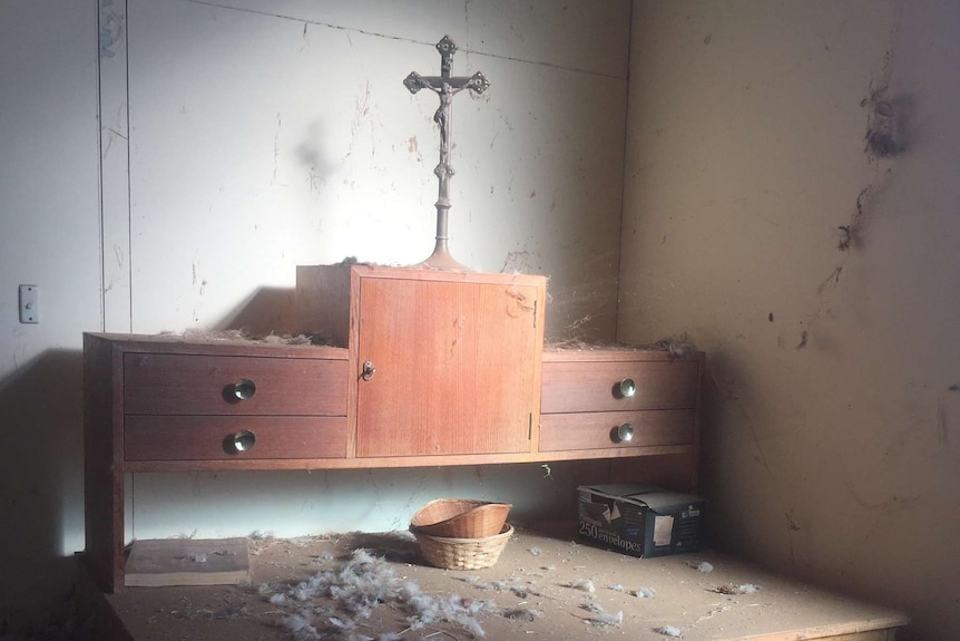 A wooden cabinet with a cross on the top sits in an old dirty room with cobwebs on the walls and feathers on the floor