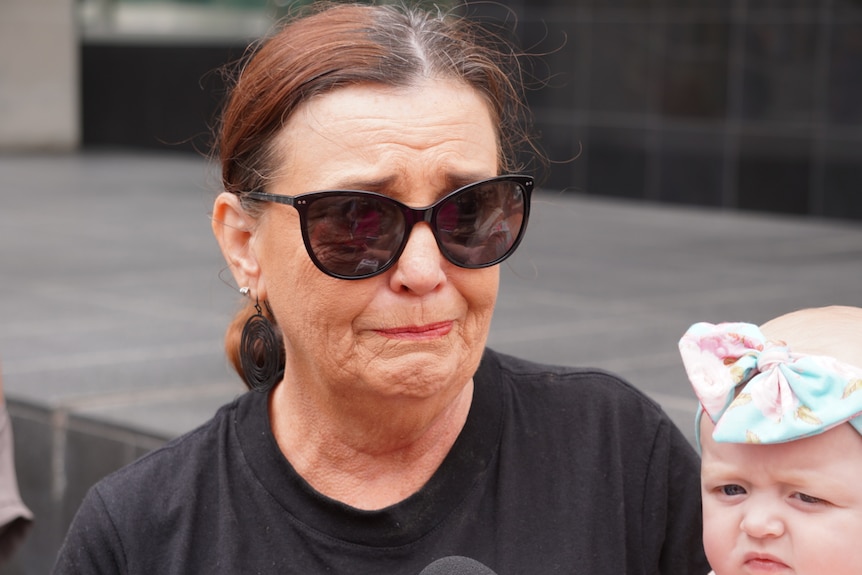 Margaret wears a black shirt and black sunglasses, holding back tears with a distraught face, while cradling a baby in her arms.