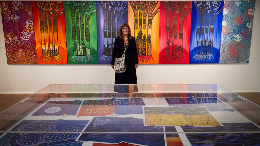 A smiling woman in a stylish black coat stands in front of a series of large, multicoloured Indigenous artworks.