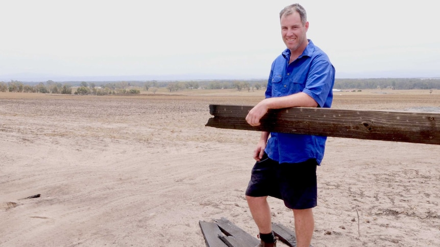 A man with a blue skirt leans on a wooden fence that burnt, with burnt pieces on the ground.