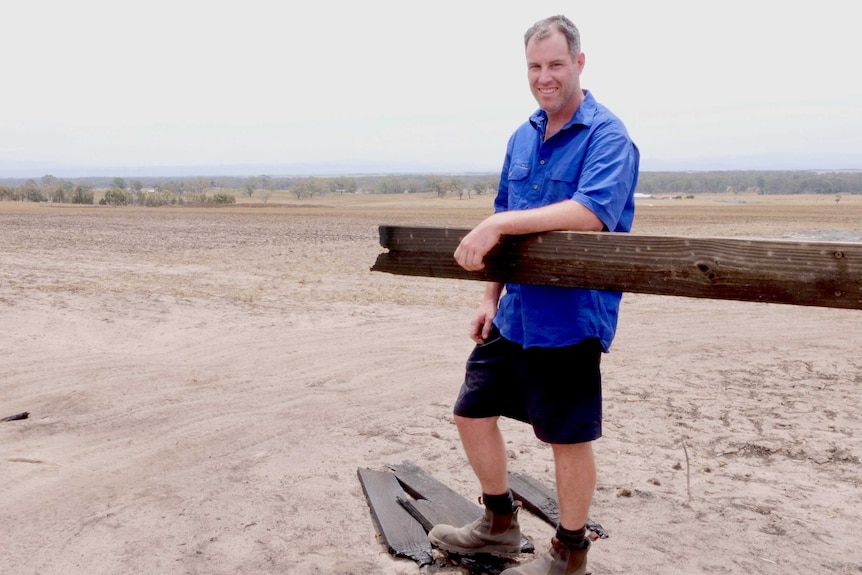 A man with a blue skirt leans on a wooden fence that burnt, with burnt pieces on the ground.