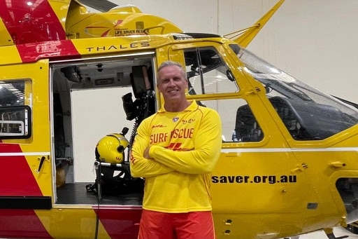 Man wearing long sleeved yellow top standing with arms folded across his chest in front of a yellow and red-striped helicopter