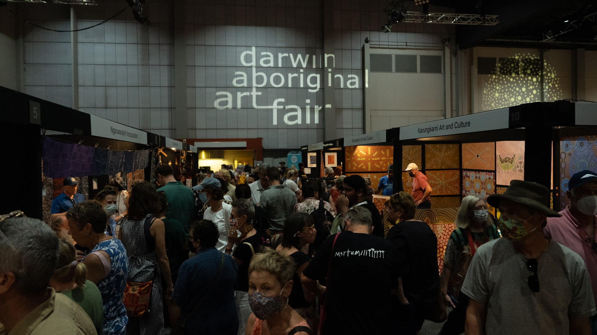 A crowd of people walking through the aisle of an art gallery, with the words "Darwin Aboriginal Art Fair" on a sign above them.