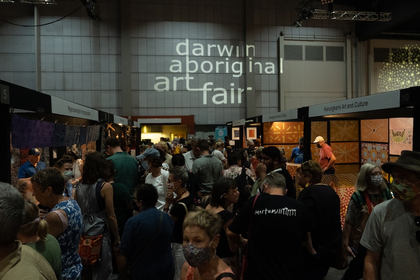 A crowd of people walking through the aisle of an art gallery, with the words "Darwin Aboriginal Art Fair" on a sign above them.