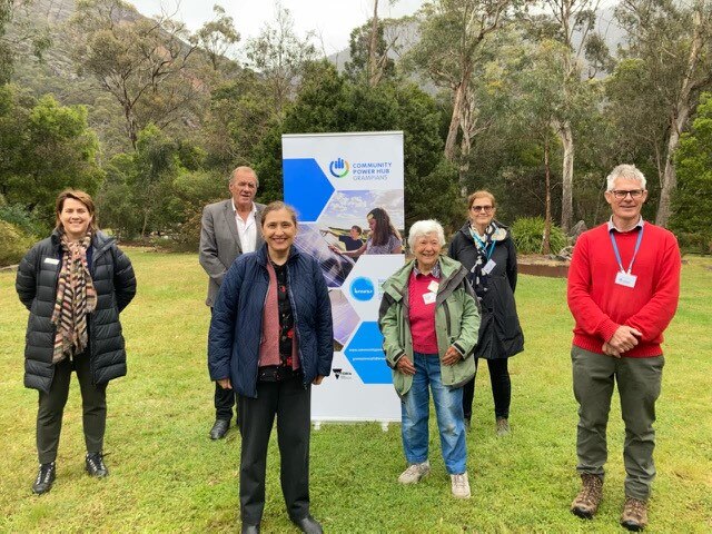 A group of people stand in front of a banner on green lawn with trees in background. 