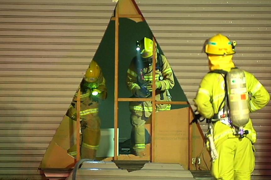 Firefighters are visible examining the scene of a fire in a factory, while firefighters watch from outside.