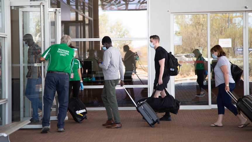 A line of people wearing masks and wheeling suitcases. They are walking through a glass door held open by a man.